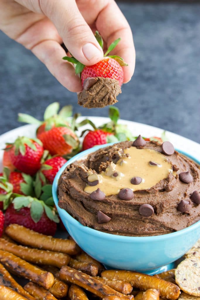 A hand dipping a strawberry into a bowl of dessert hummus with a peanut butter drizzle. 