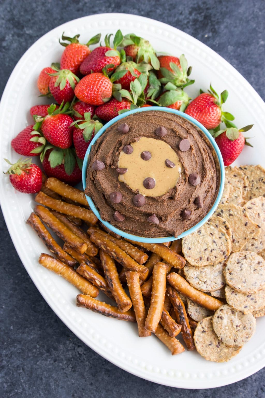 A bowl of chocolate peanut butter hummus on a white platter with fruit and crackers around it.