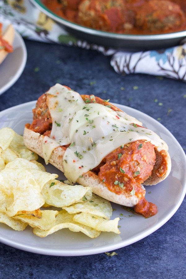 A vegan meatball marinara sub with melted cheese on top next to chips on a gray plate with a dark blue background.