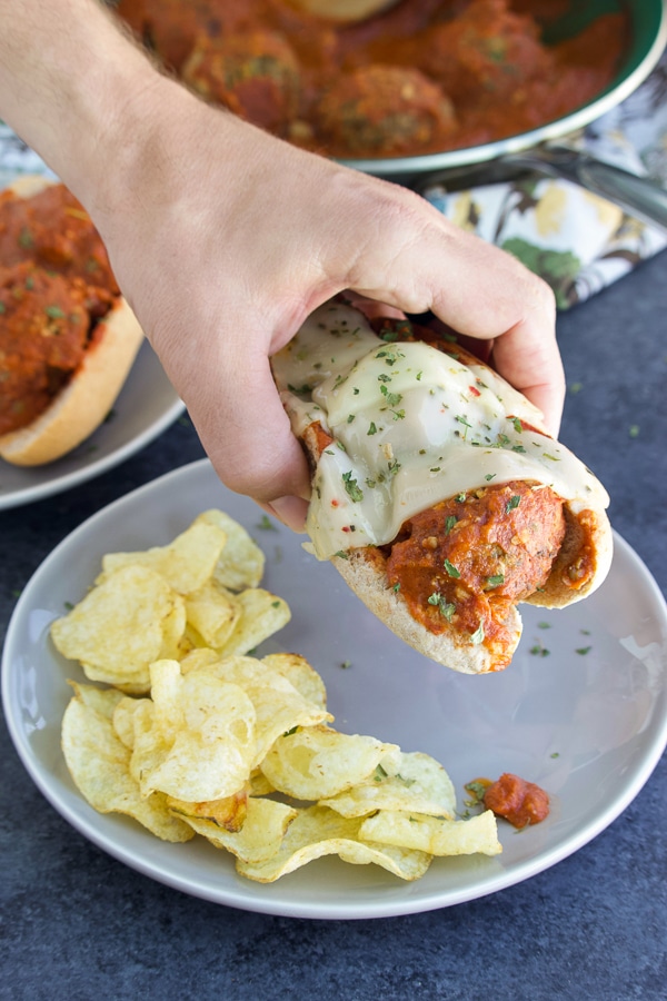 A hand picking up a sub from a gray plate with chips on a gray background.