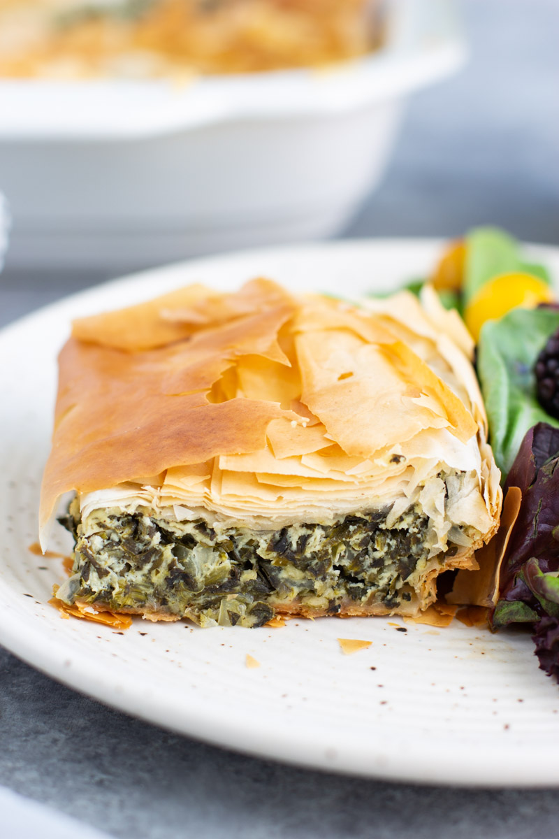 A piece of spinach pie on a white plate with a salad on a gray background. 