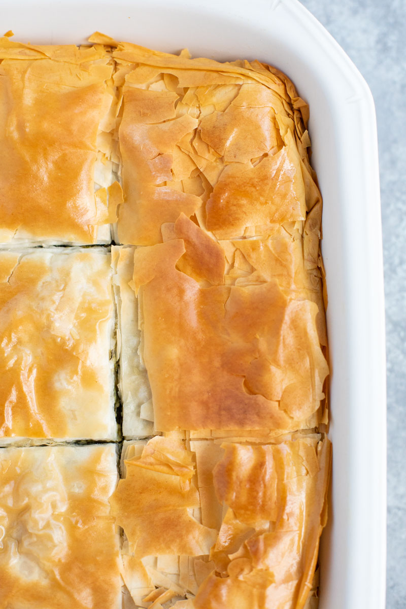 A close up view of the spinach pie cut into squares with crispy filo topping in a white casserole dish.