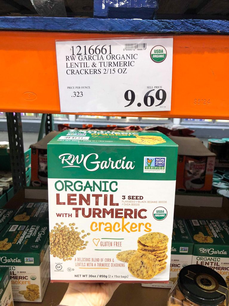 A hand holding a box of organic vegan RW Garcia lentil turmeric crackers for $9.69 at Costco.