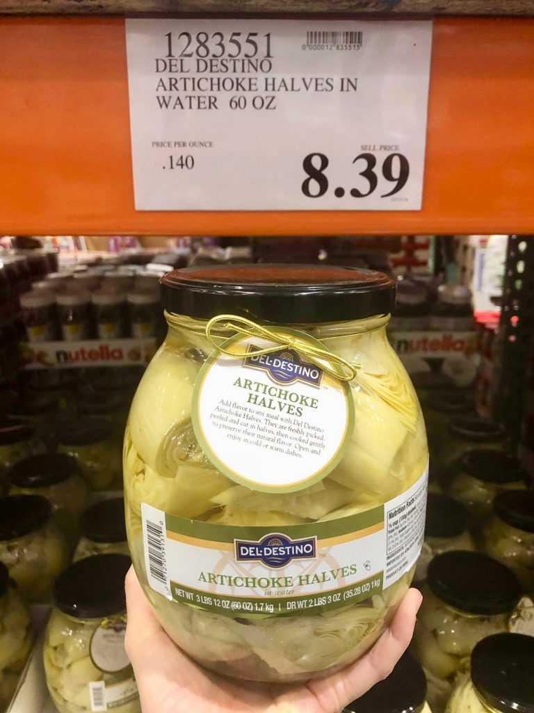 A hand holding a glass jar of vegan artichoke halves for $8.39 at Costco.