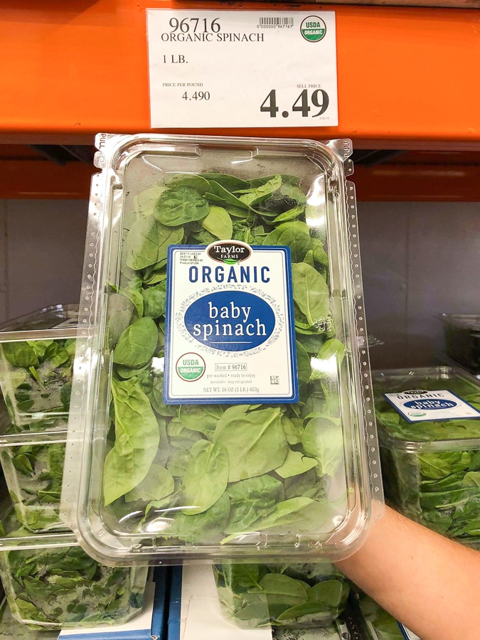 A hand holding a container of organic baby spinach for $4.49 at Costco.