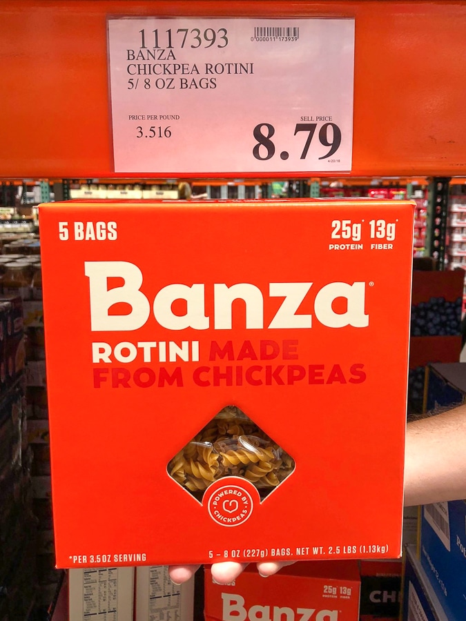 A hand holding a red box of vegan Banza pasta for $8.79 at Costco. 