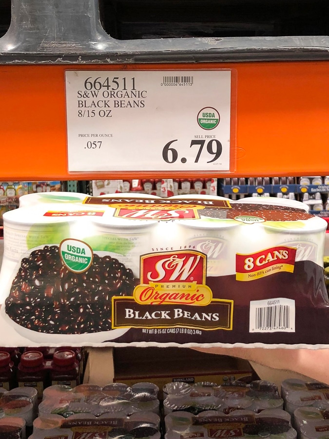 A hand holding an 8-pack of organic vegan canned black beans for $6.79 at Costco.