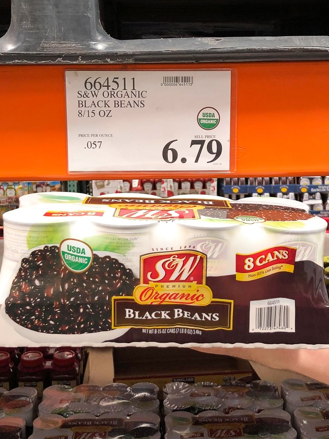 A hand holding an 8-pack of organic vegan canned black beans for $6.79 at Costco.