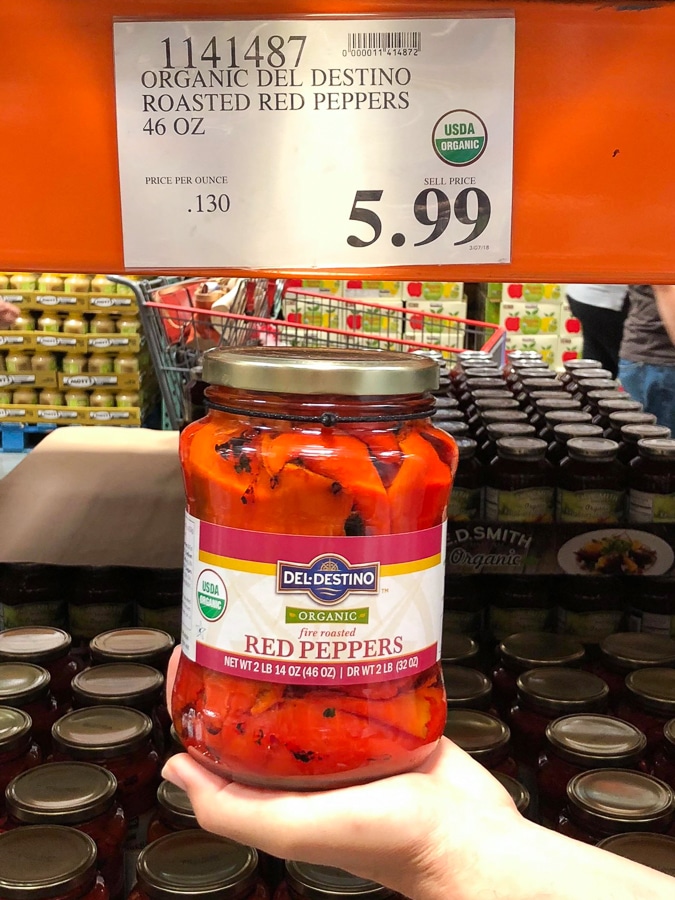 A hand holding a glass jar of fire-roasted organic vegan red peppers for $5.99 at Costco. 