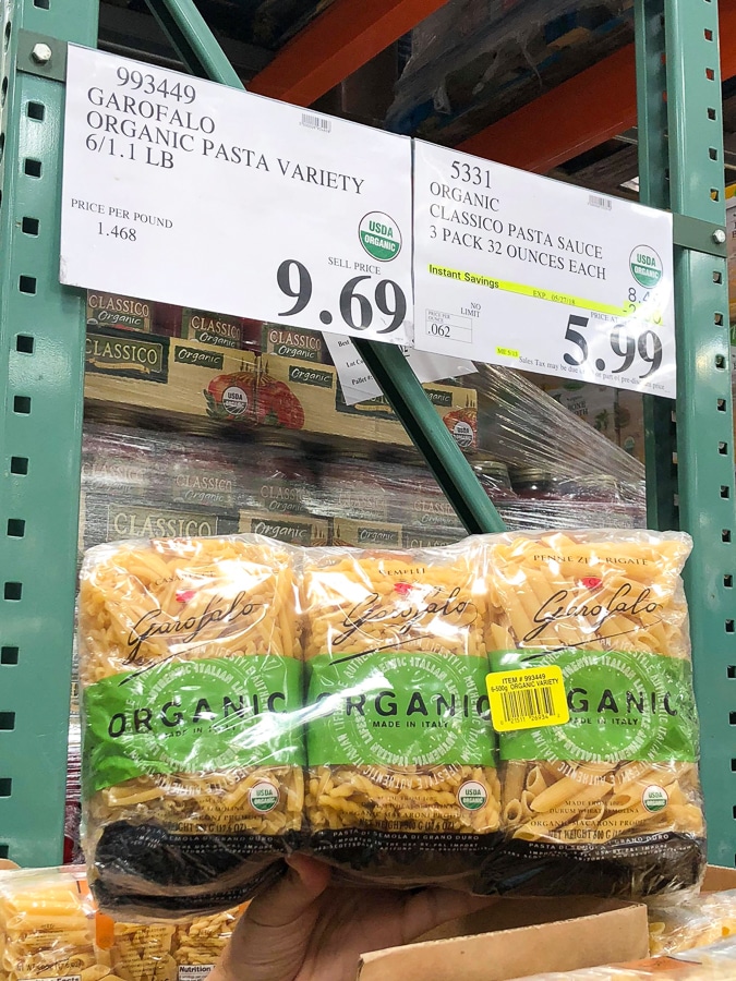 A hand holding a 6-pack of organic vegan pasta for $5.99 at Costco.