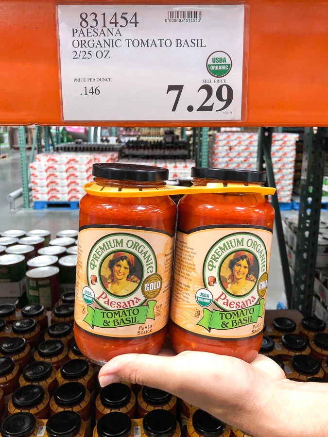 A hand holding a 2-pack of organic vegan tomato basil sauce in glass jars for $7.29 at Costco.