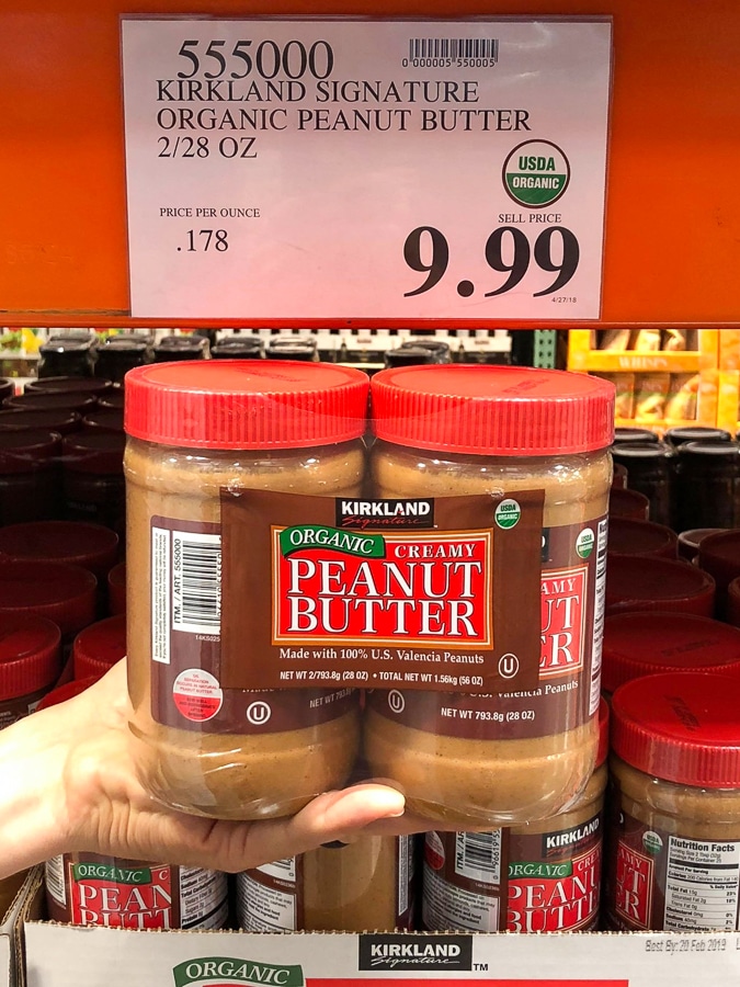 A hand holding a 2-pack container of organic vegan peanut butter for $9.99 at Costco.