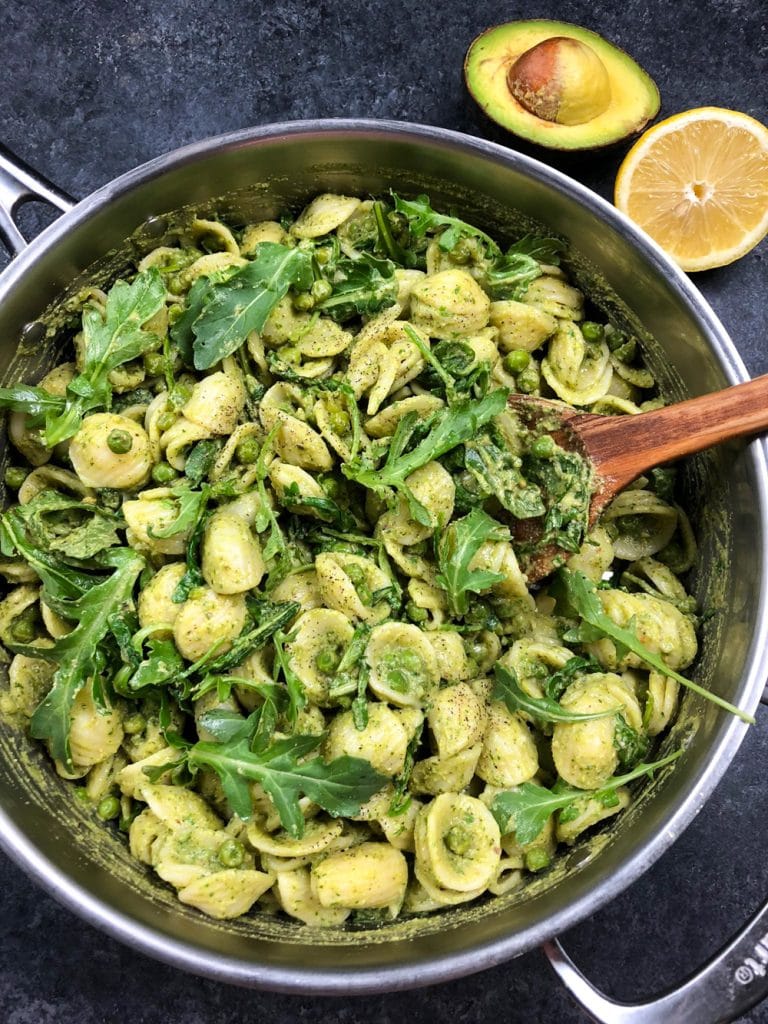 A large pan full of pasta tossed in vegan pesto sauce and mixed with arugula and green peas on a dark background. 
