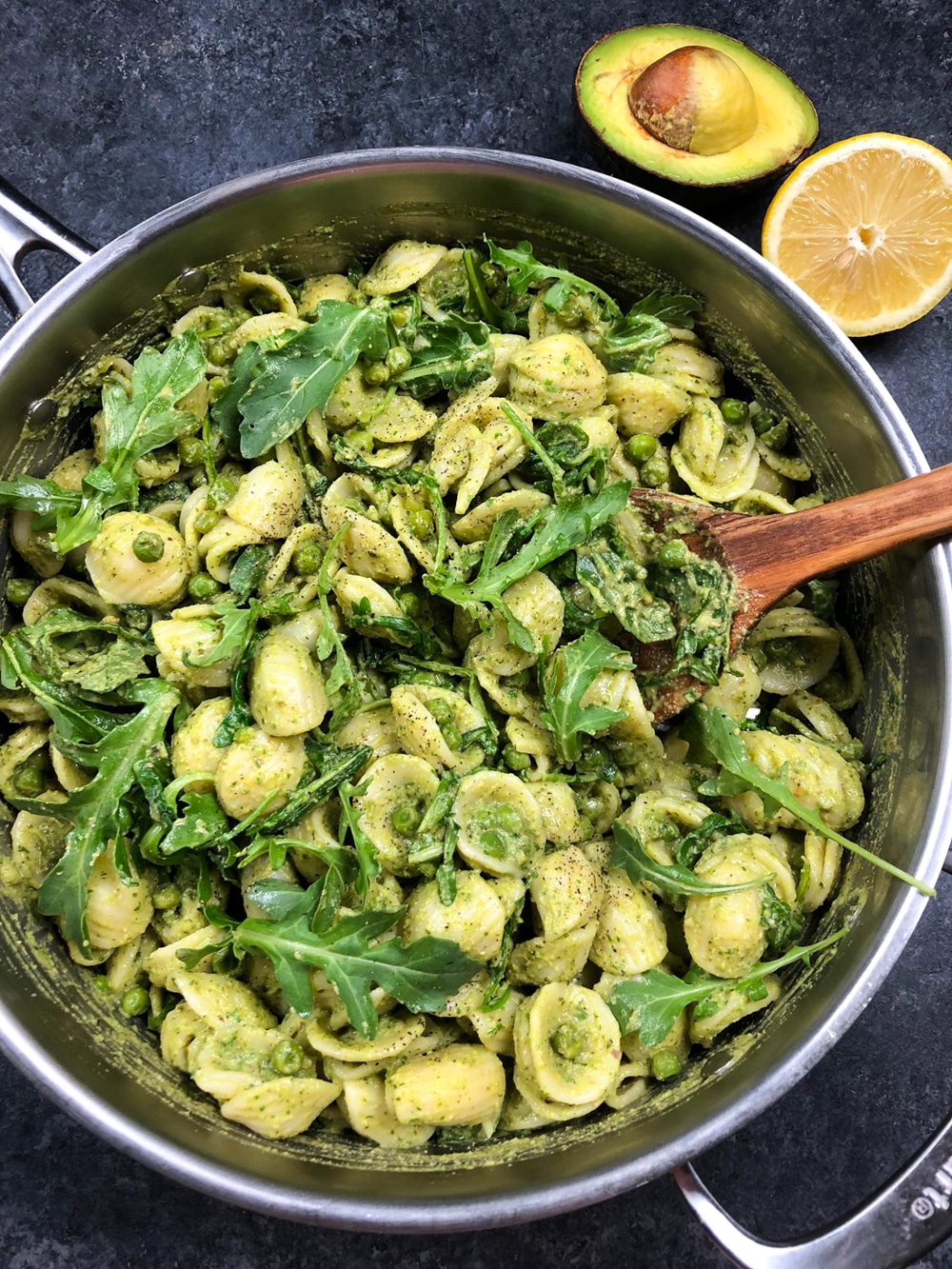 A large pan full of pasta tossed in pesto sauce and mixed with arugula and green peas on a dark background. 