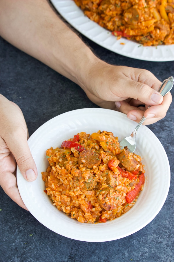 Two hands holding a white bowl full of vegan sausage, peppers, and rice on a dark textured background.