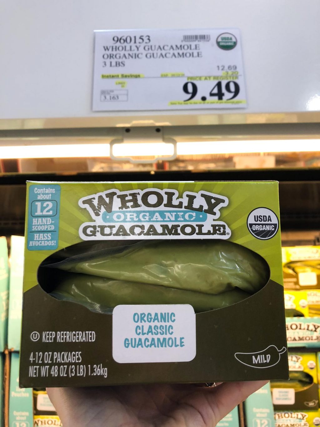 A hand holding a box of organic vegan Wholly Guacamole for $9.49 at Costco.