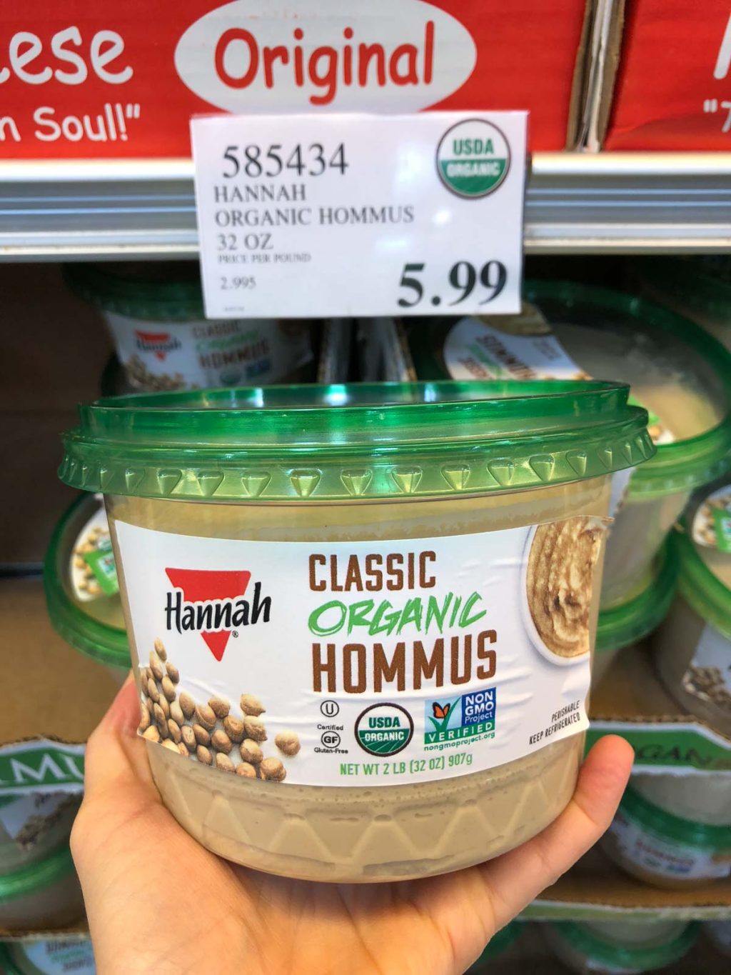 A hand holding a container of organic vegan hummus for $5.99 at Costco.