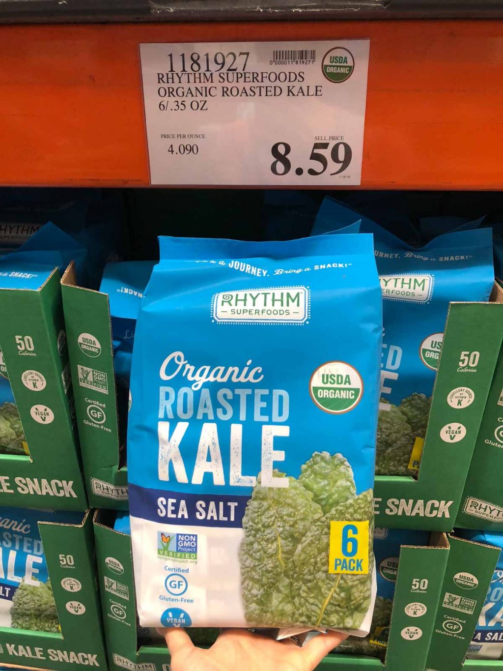 A hand holding a blue container of Rhythm organic vegan roasted kale snacks for $8.59 at Costco.