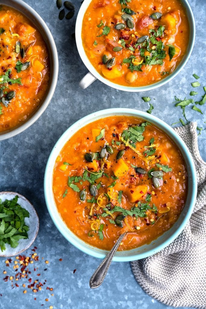 Three garnished bowls of sweet potato lentil soup on a blue textured background. 