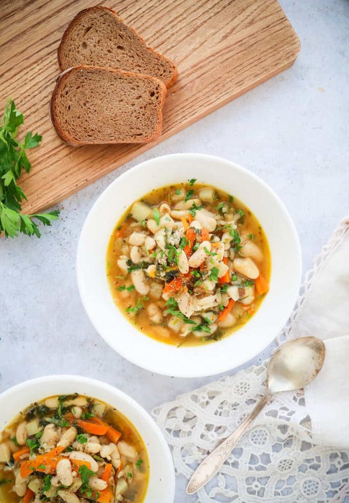 Two white bowls of vegan white bean soup on a light background next to a cutting board. 