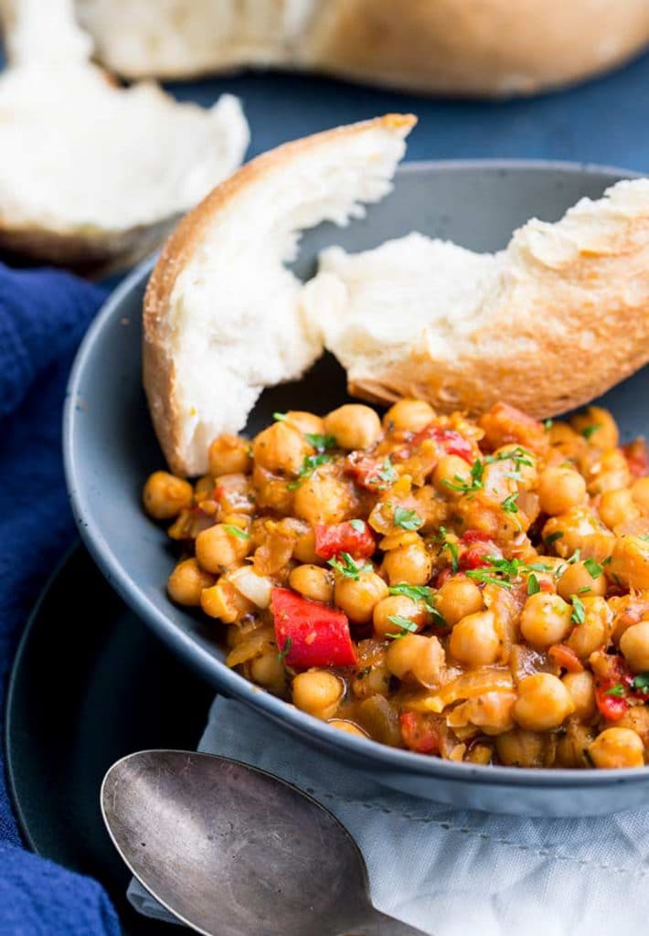 A blue bowl of tomato and chickpea stew with a ripped piece of bread on a blue background. 