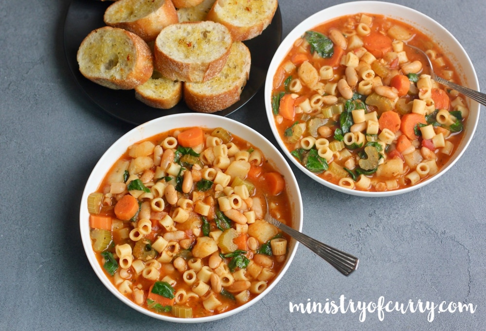 Two white bowls of pasta e fagioli next to a plate of bread sitting on a grey background. 