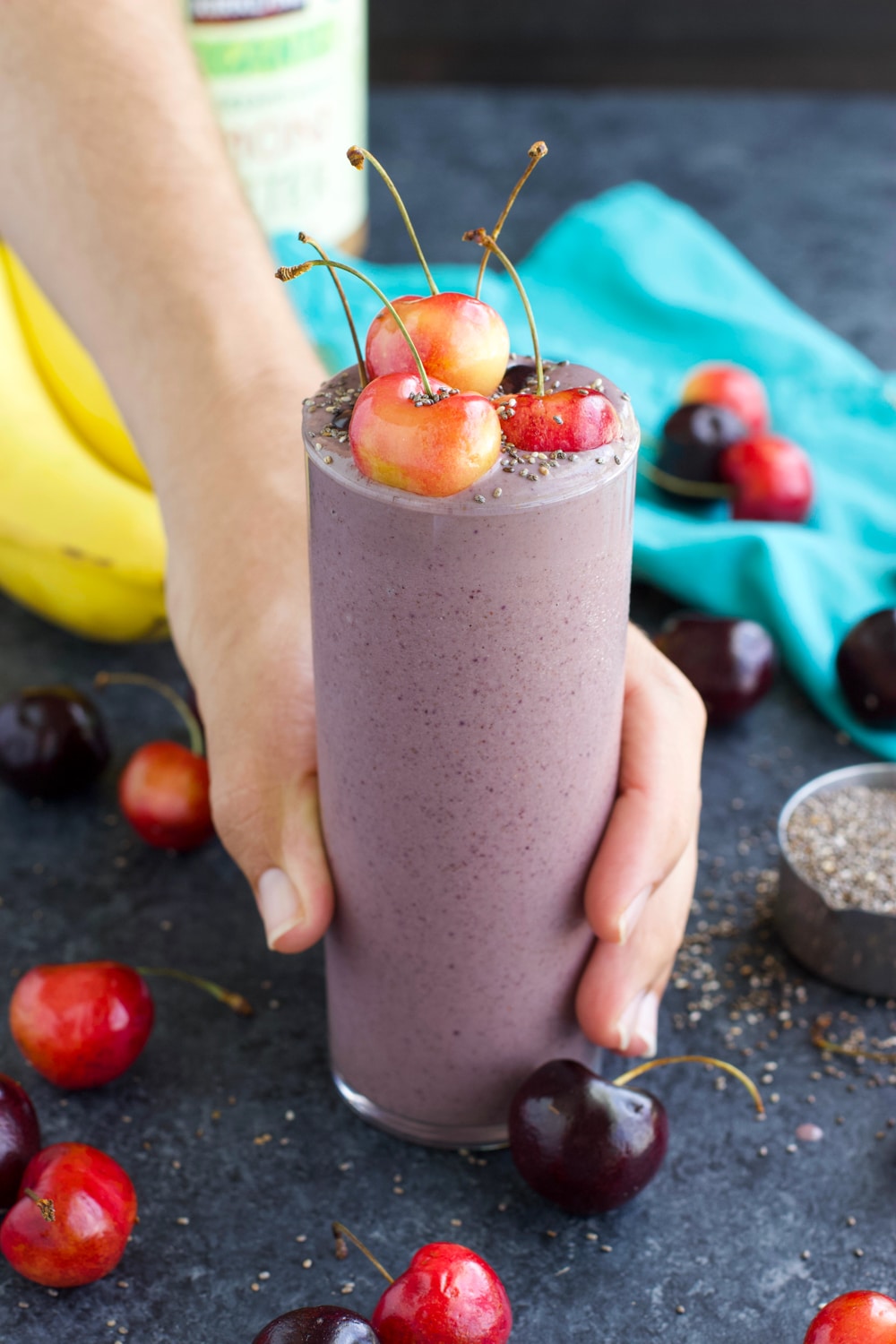 A hand holding a smoothie topped with cherries in a glass cup on a dark background.