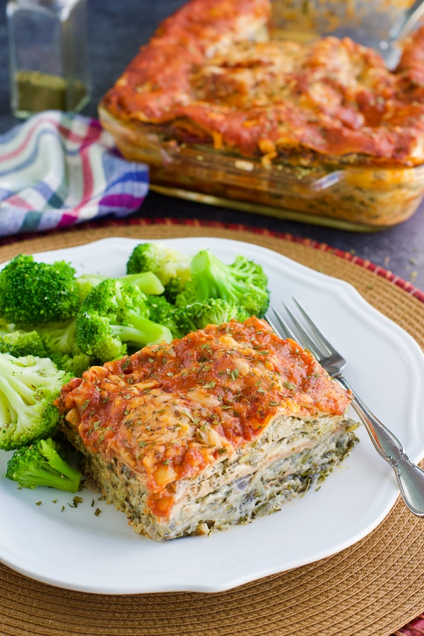 A piece of vegan lasagna and a side of broccoli on a white plate next to a tray of lasagna.