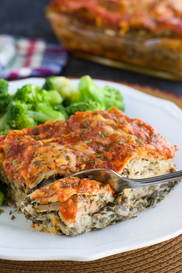 A fork cutting a piece from a slice of lasagna next to broccoli on a white plate.