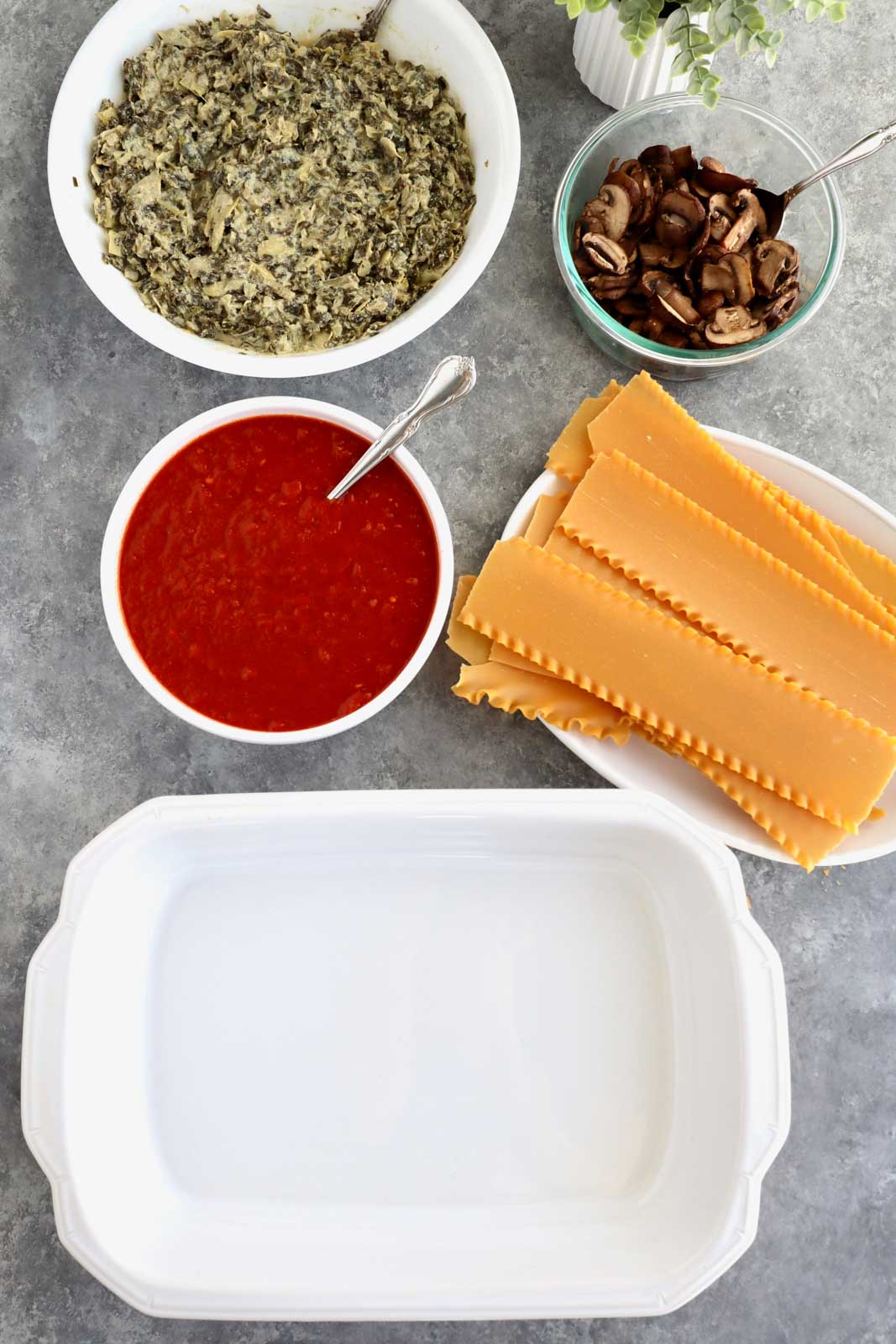 A white casserole dish, lasagna noodles, tomato sauce, mushrooms, and spinach dip in bowls.