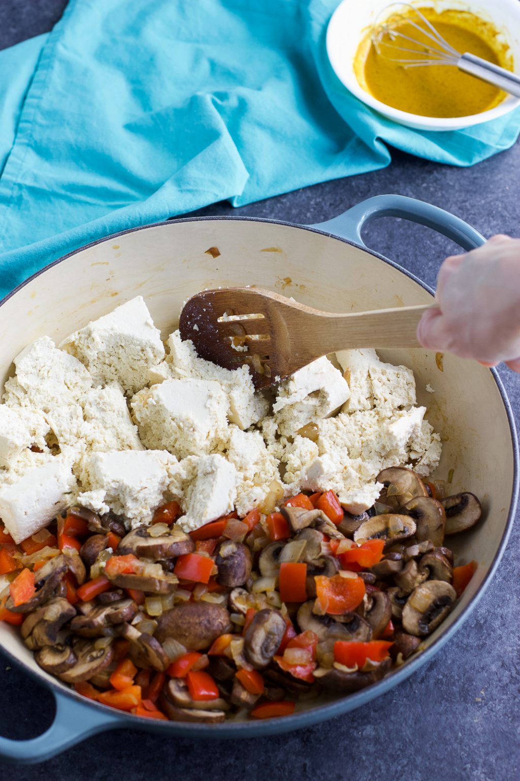 A hand stirring crumbled tofu and vegetables in a large blue pan next to a small bowl on a dark background.