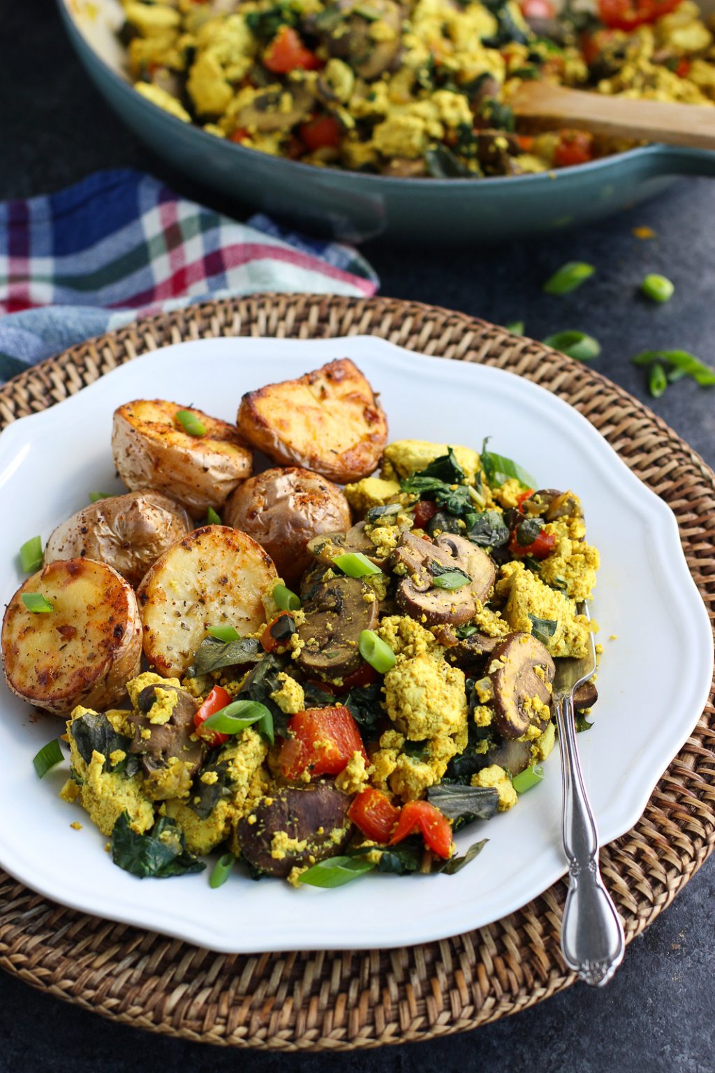 Tofu scramble on a white plate with potatoes on top of a wicker charger next to a blue pan on a dark background.