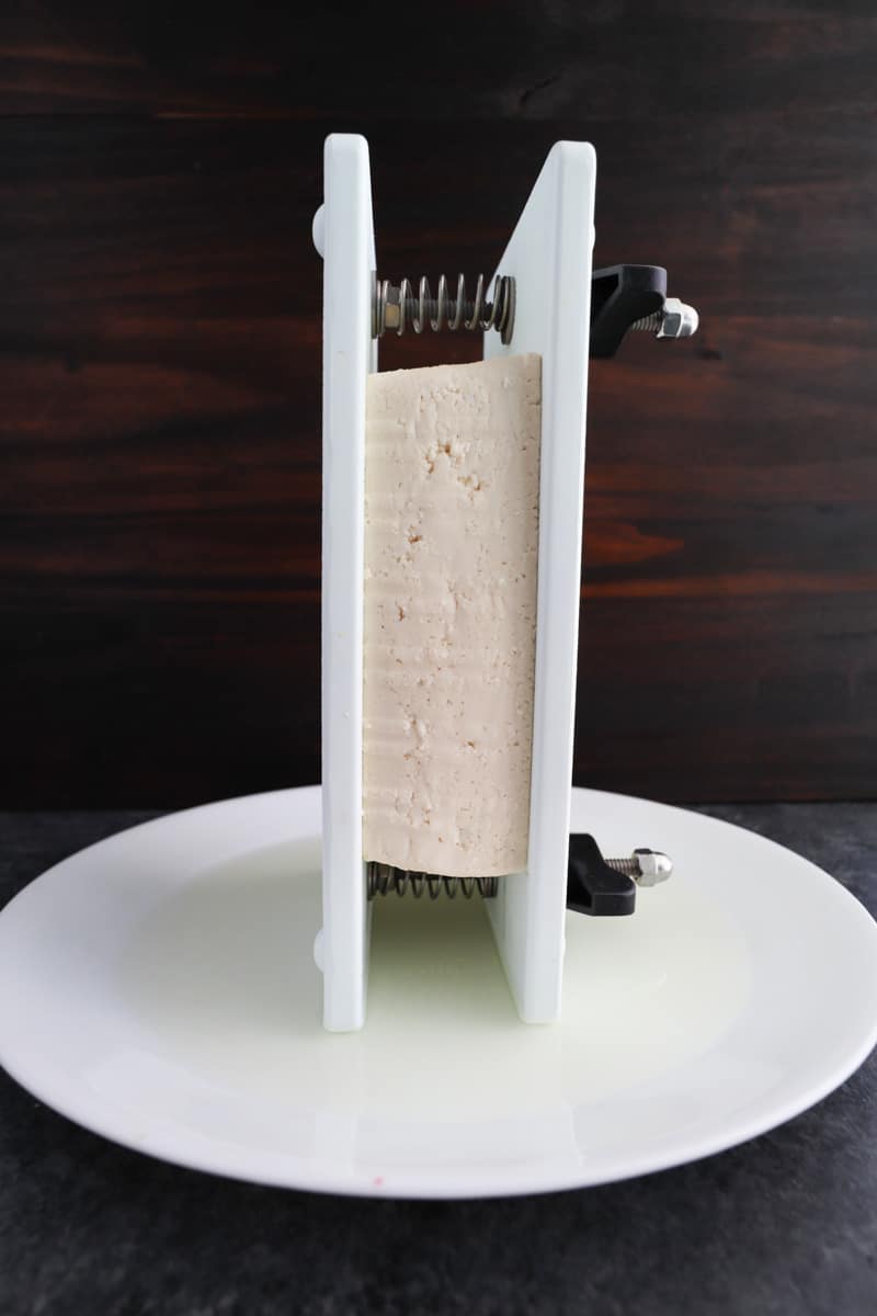 A block of tofu in a press being drained on top of a white plate on a dark background. 