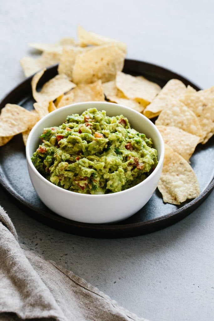 A small white bowl of vegan keto guacamole is on top of a matte black plate that is holding tortilla chips and it's resting on a neutral background..