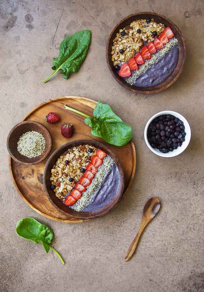 Two vegan keto breakfast blueberry smoothie bowls topped with hemp seeds, strawberries, and granola on a wooden platter that is resting on a neutral background.