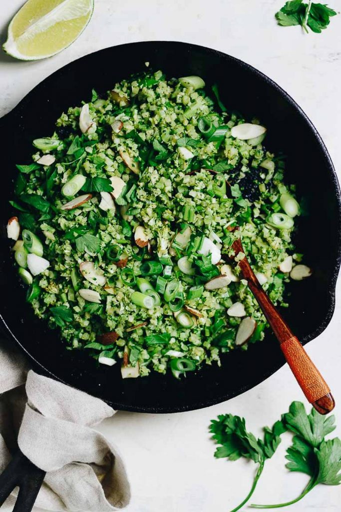 A low-carb vegan side dish of broccoli fried rice with fresh herbs, green onions, and sliced almonds in a cast iron skillet with a white background.