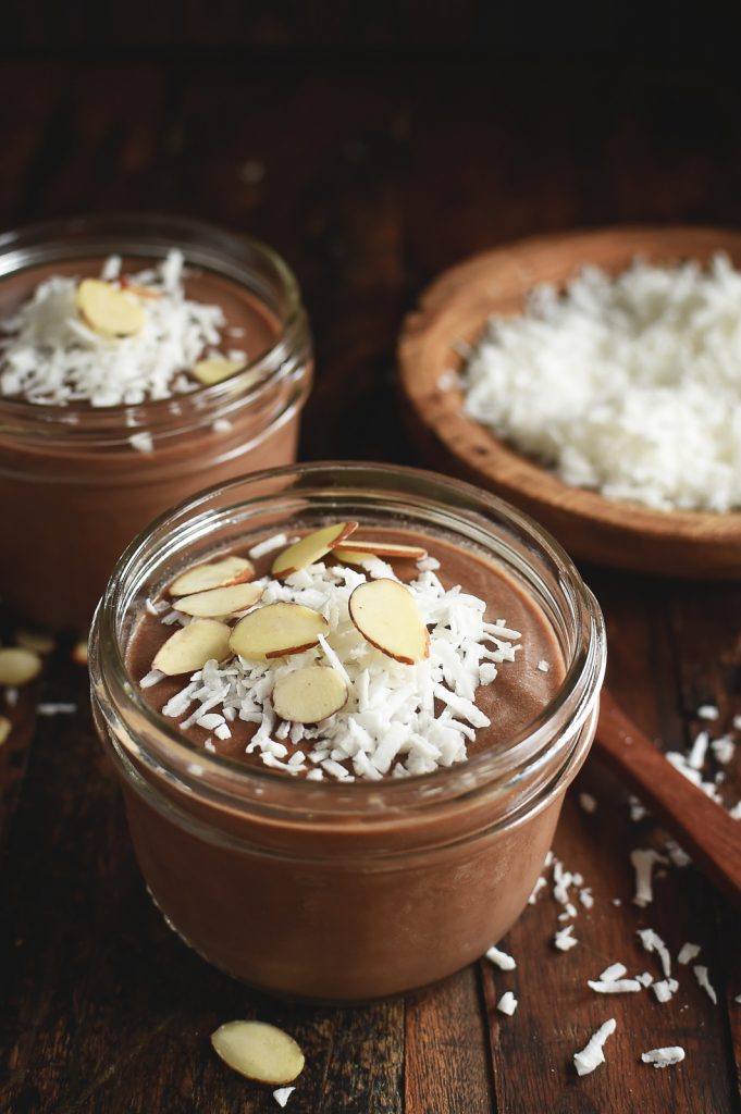 A vegan keto dessert of chocolate almond pudding in two glass jars with a bowl of coconut to the right on top of a rustic background.