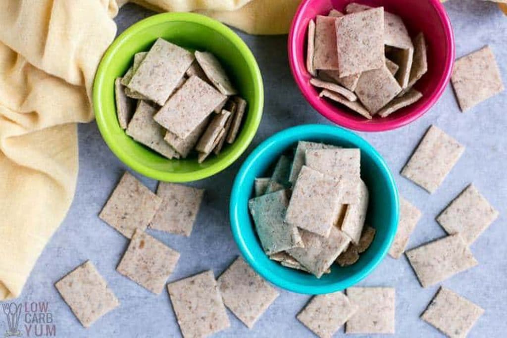 Three multi-colored bowls hold low-carb vegan keto crackers on a gray background with a soft yellow towel on the side.