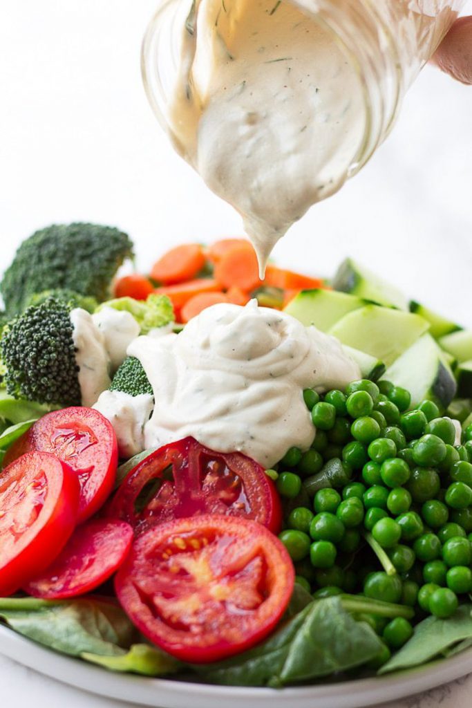 A hand pouring a glass jar of vegan keto ranch dressing over a vegetable loaded plate of salad on a white background.