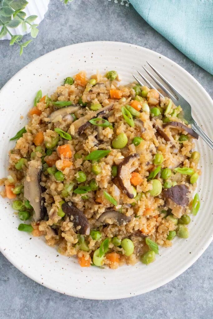 A vegan keto dinner featuring low-carb cauliflower fried rice with broccoli, carrots, and cilantro on a texture white plate with a rustic background.