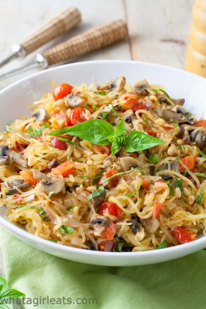 Vegan keto pasta with spaghetti squash, mushrooms, tomatoes, and fresh basil served in a white bowl on top of a white table with a green napkin on the side.