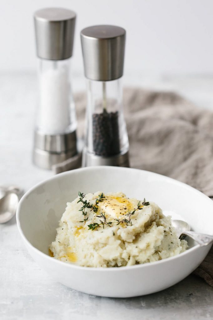 A white bowl filled with mashed cauliflower with salt and pepper grinders next to it on a white background.