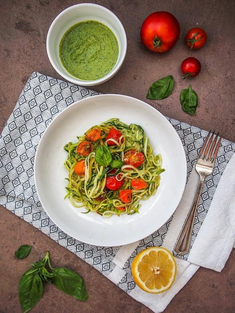 A large white bowl filled with vegan keto zucchini noodles, pesto sauce, and tomatoes with a patterned napkin underneath is and pesto ingredients scattered around.