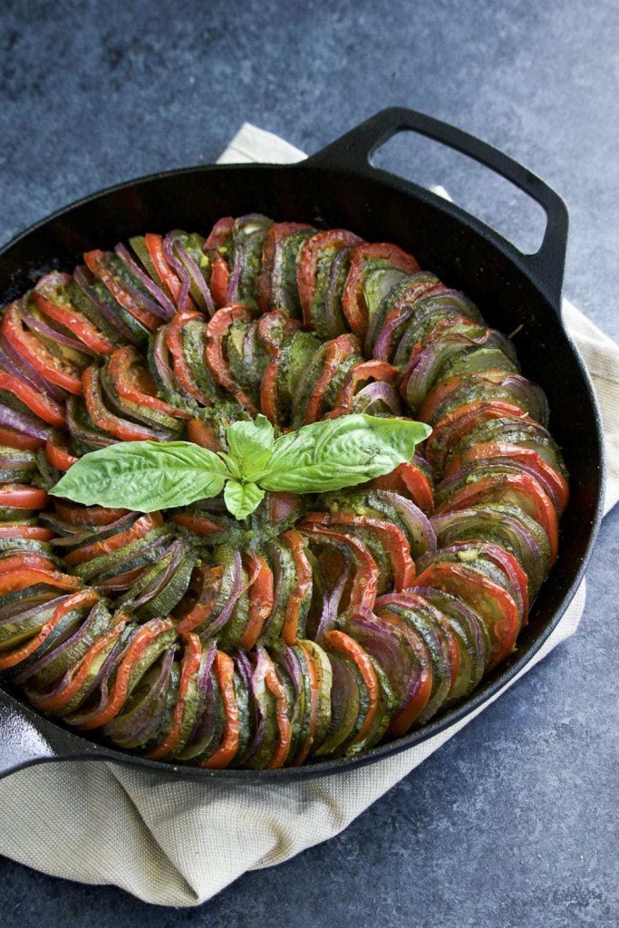 A cast iron skillet filled with thinly sliced zucchini, tomatoes, red onions, and pesto sauce resting on a nude towel with a blue background.