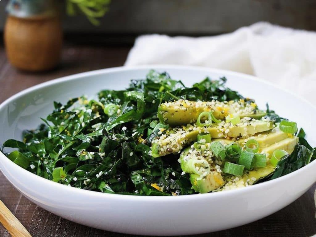 A vegan keto salad with three types of greens and avocado, hemp seeds, and green onions is served in a white bowl on a rustic background. 