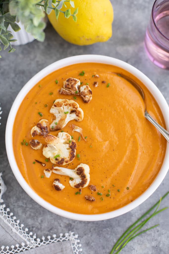 A bowl of vegan keto soup made of cauliflower, coconut milk, and red curry paste is topped with green onions and hot sauce in a flower bowl and it's sitting on a rustic cutting board.