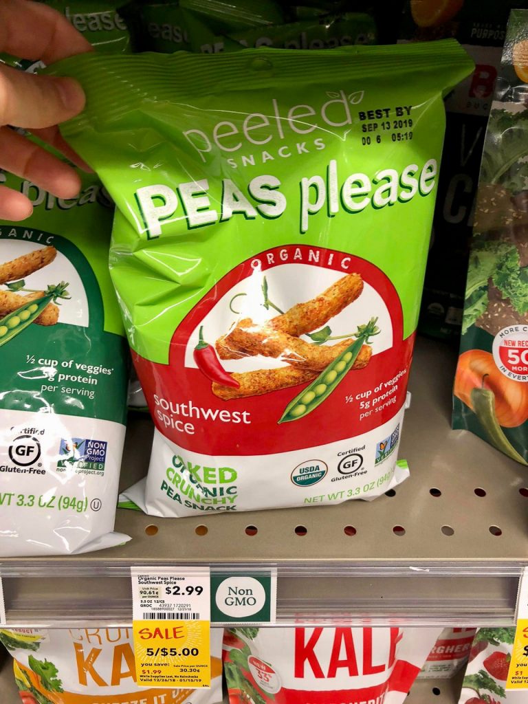 A hand holding a bag of vegan peas please snacks on a shelf at Whole Foods. 