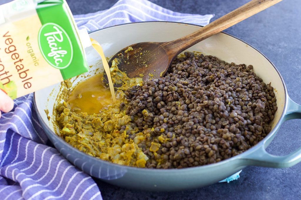 A large pan filled with lentils, curry paste, a wooden spoon and a hand pouring vegetable broth into the pan. 