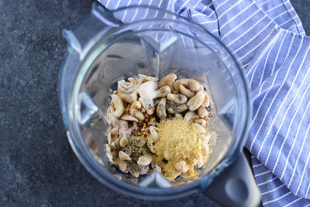 A large blender cup filled with cashews and seasonings next to a blue napkin. 