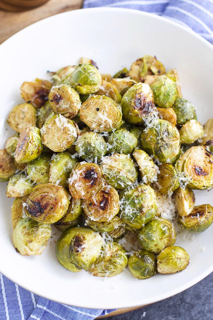 Low-carb vegan roasted brussel sprouts with garlic and vegan parmesan cheese on a white platter with a blue and white striped dish towel under it. 